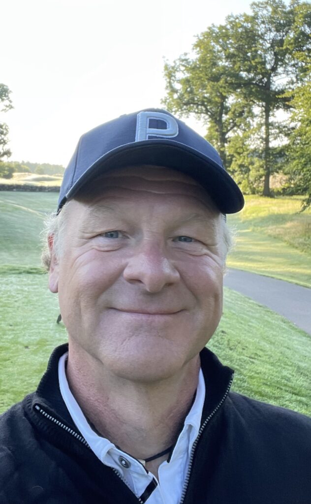 A man wearing a baseball cap and casual outdoor clothing, smiling at the golf course with trees in the background.